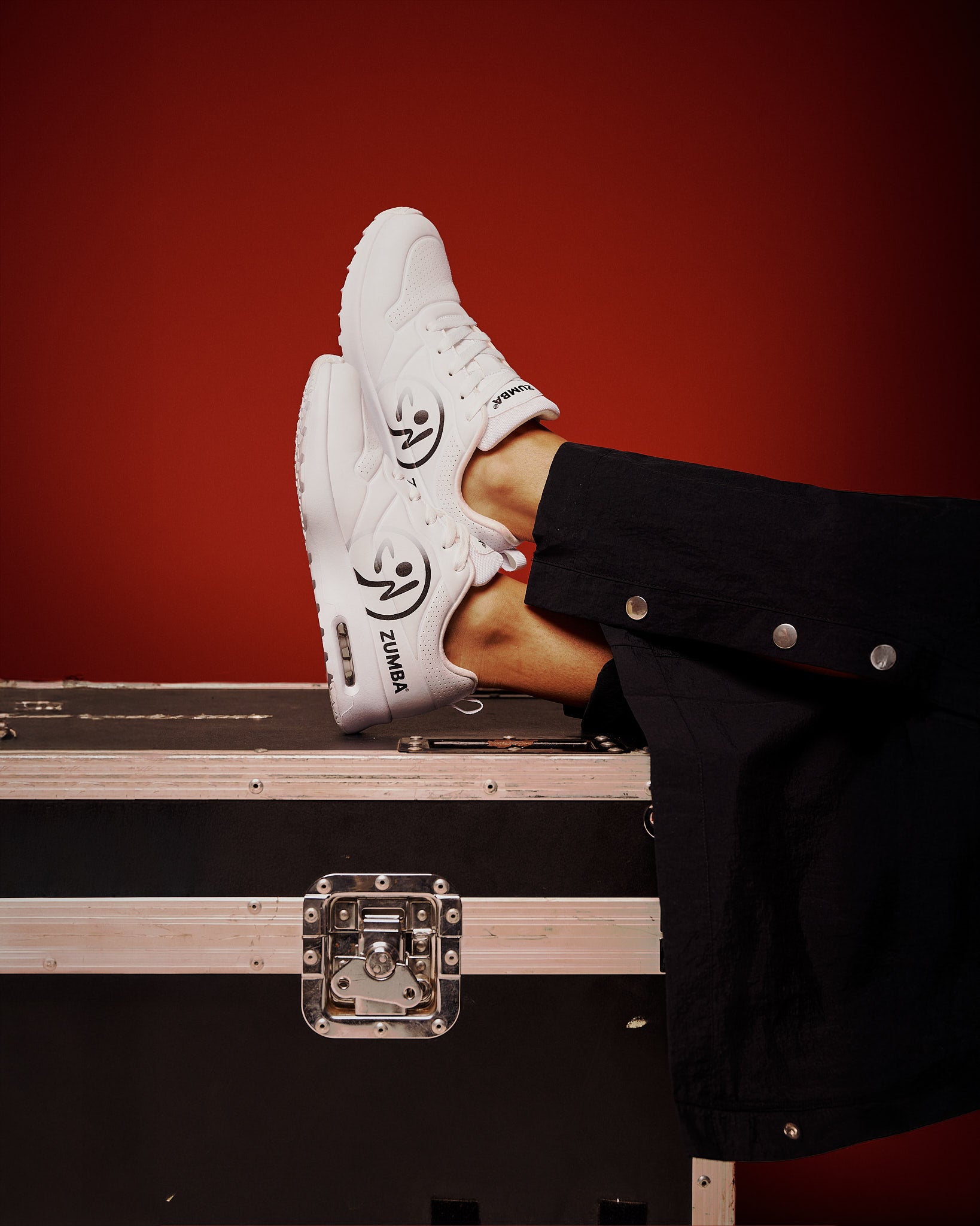 A pair of white low-top sneakers with black graphic details and the brand name 'ZUMBA' visible on the side, worn with black pants and posed on top of a black equipment case against a dark red background.