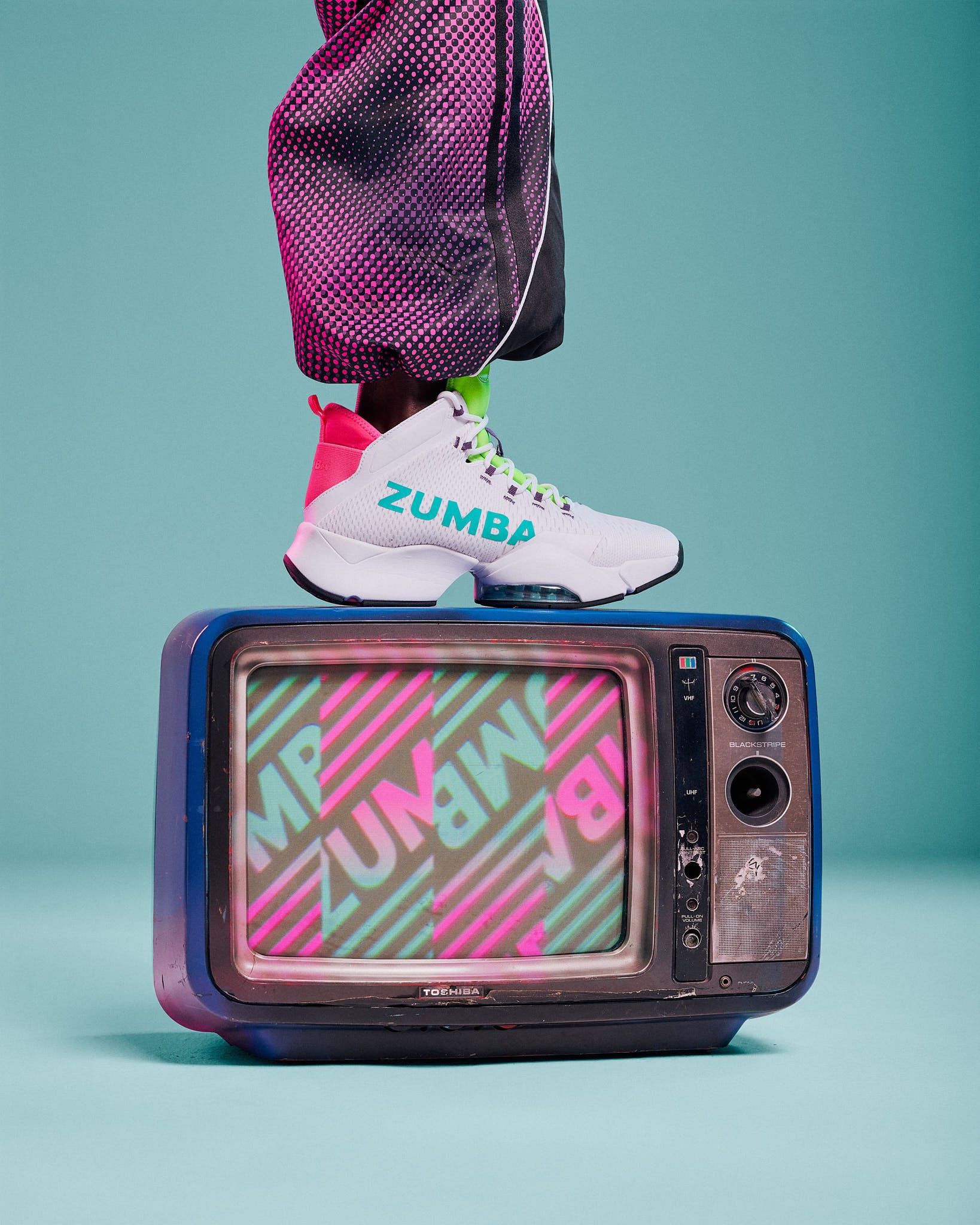 White high-top sneaker with aqua "ZUMBA" text and pink and green accents, propped on a vintage TV against an aqua background.