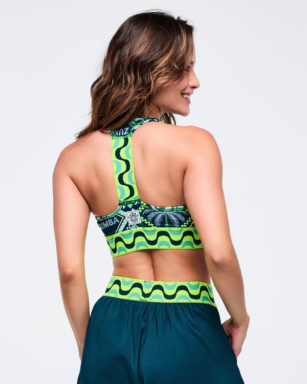 Back view of the green printed Zumba sports bra showing the racerback strap and lime wave-print band, paired with teal jogger pants with a matching wave waistband. Female model is shown on a light gray studio background.
