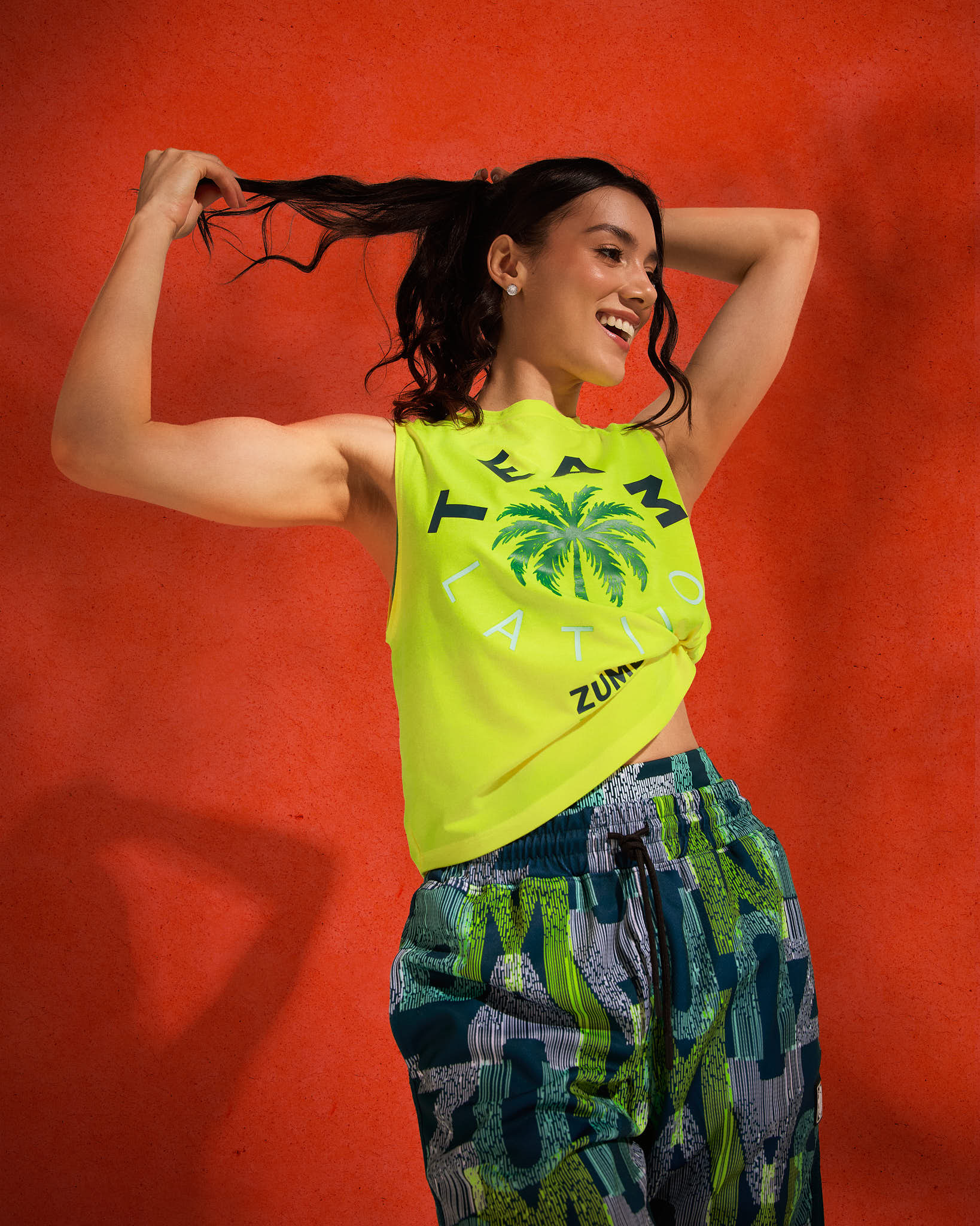 Neon yellow “TEAM LATINO” sleeveless Zumba tank, knotted at the waist, paired with matching green-and-black abstract print jogger pants; female model poses with arms raised against a red backdrop.