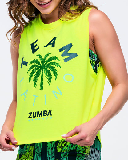 Neon yellow sleeveless “TEAM LATINO” Zumba tank with palm tree graphic and bold lettering; female model wears green-and-black abstract print biker shorts, holding the hem to show the relaxed fit on a white background.