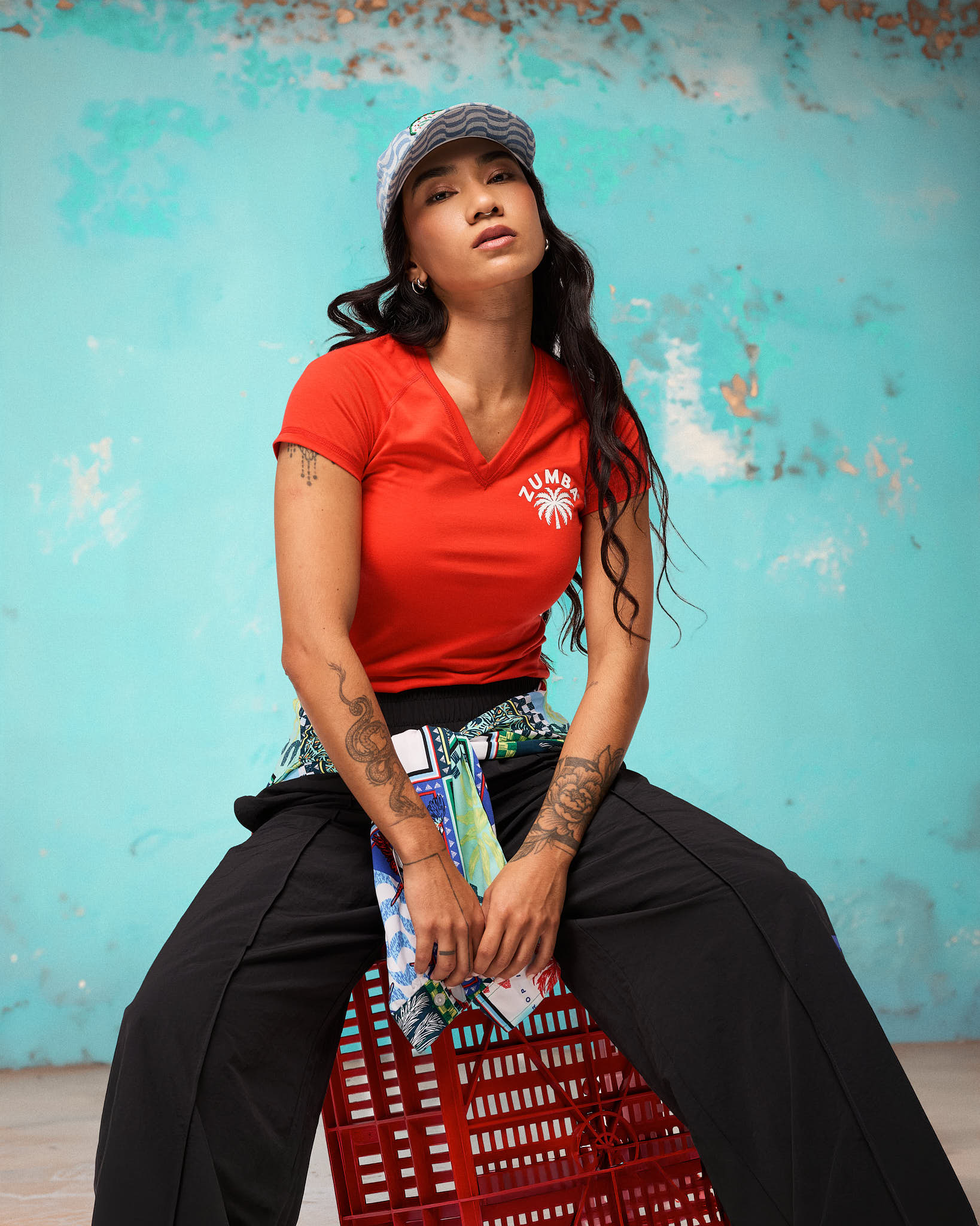Red fitted V-neck Zumba tee with small arched ZUMBA above a white palm logo on the left chest. Female model is also wearing loose black wide-leg pants, a colorful graphic jacket tied at the waist, and a blue-and-white patterned cap; she sits on a red crate against a turquoise wall.