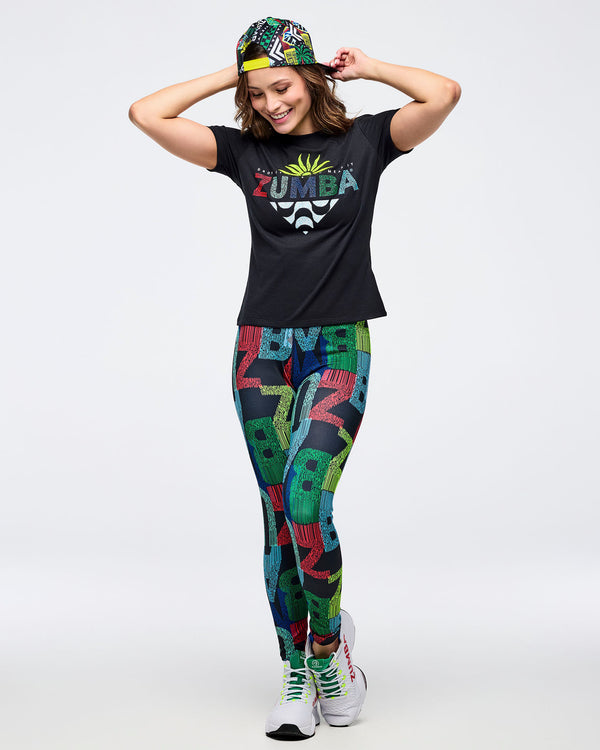 Black “ZUMBA” graphic tee with multicolor logo and palm graphic, paired with high-waisted, multicolor letter-print leggings; female model wears a colorful patterned snapback cap backward and white sneakers with white laces with neon green eyelets, posing with hands behind head on a white backdrop.