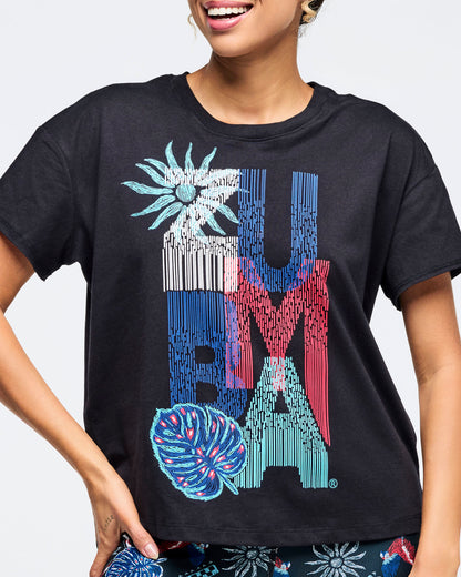 Close-up of a black short-sleeve crewneck tee with oversized “ZUMBA” vertical graphic in blue, red, teal and white with tropical flower and leaf accents; female model shown from chest to hips wearing matching dark tropical-print leggings on a white background.