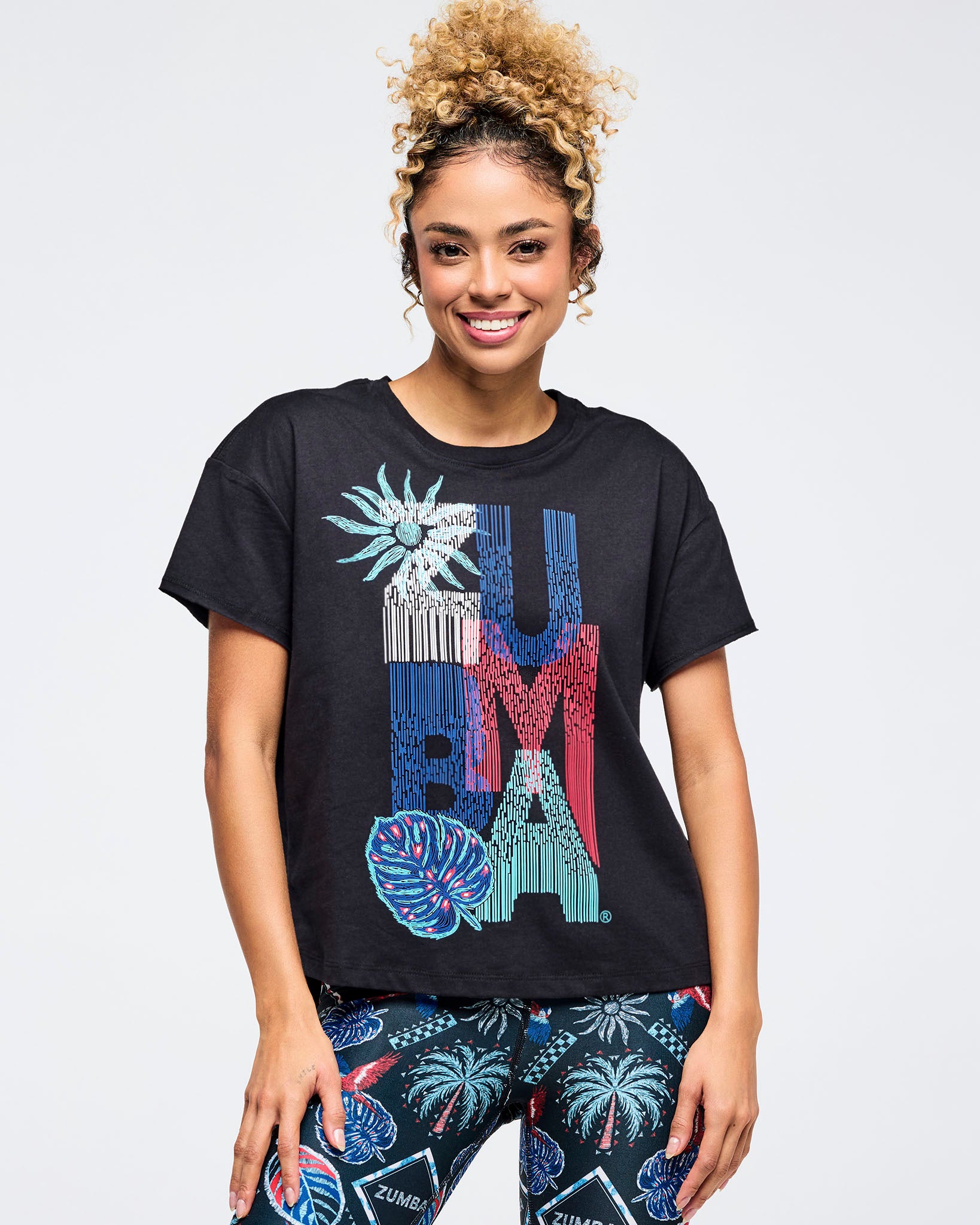 Front view of a black relaxed-fit crewneck “ZUMBA” graphic tee with colorful tropical-inspired artwork, worn with dark tropical-print leggings on a smiling female model against a white background.