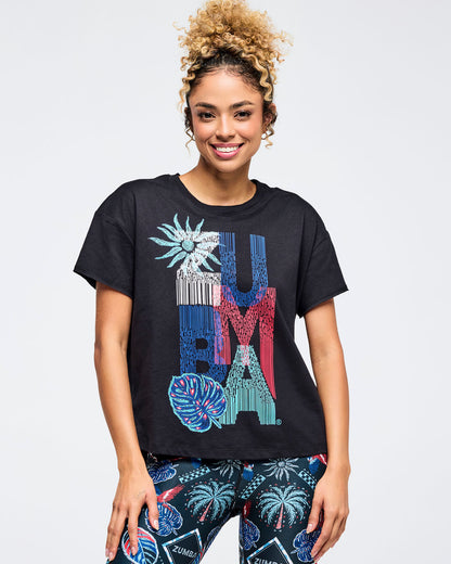 Front view of a black relaxed-fit crewneck “ZUMBA” graphic tee with colorful tropical-inspired artwork, worn with dark tropical-print leggings on a smiling female model against a white background.