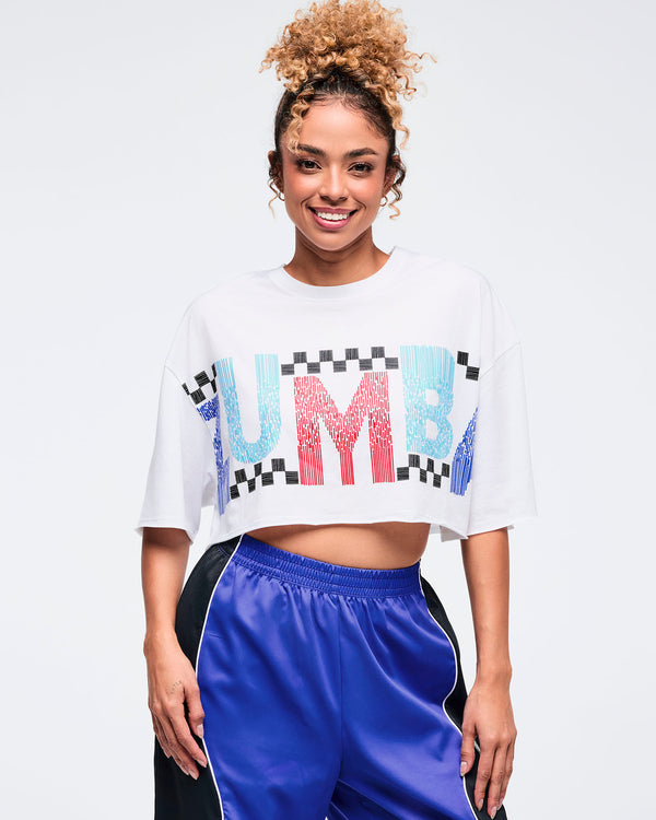White cropped oversized T-shirt with multicolor “ZUMBA” graphic and checkered details, worn by a female model with shiny royal blue track pants and white athletic sneakers.