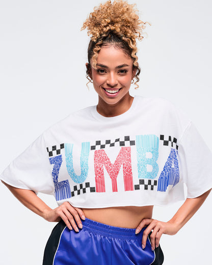 White cropped oversized T-shirt with bold multicolor “ZUMBA” graphic and black checkered accents across the chest, worn by a female model with shiny royal blue track pants featuring black side panels and white piping.