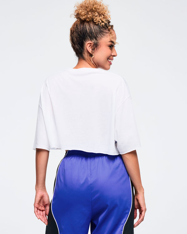 Back view of the white cropped oversized T-shirt on a female model, showing the loose sleeves and straight cropped hem, paired with shiny royal blue track pants with black side panels and white piping.