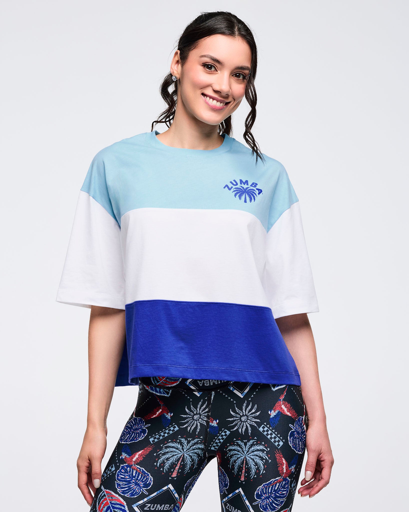 Color-block oversized T-shirt in light blue, white, and royal blue with a small Zumba palm logo on the chest, worn by a female model with black tropical-print leggings.