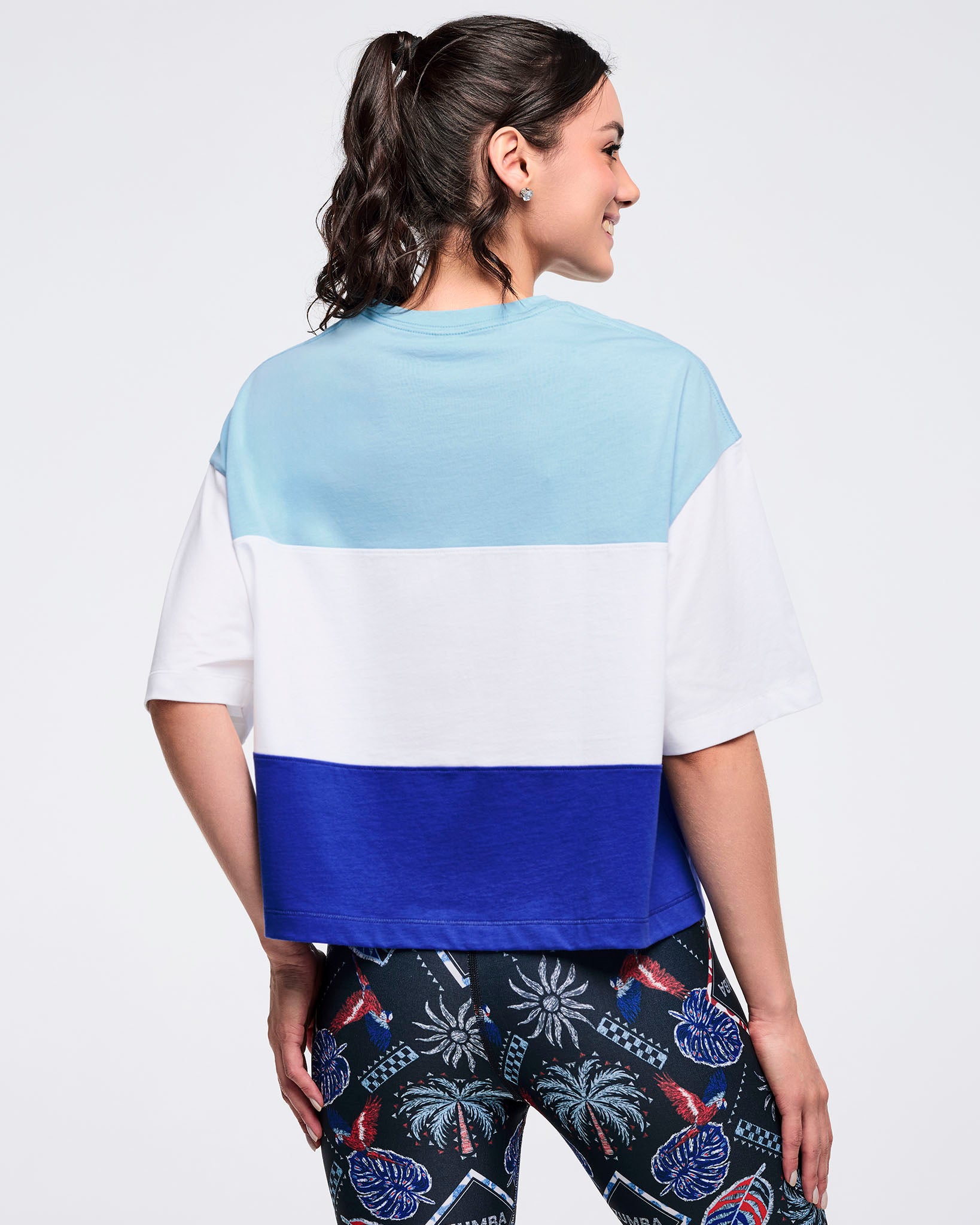 Back view of the light blue, white, and royal blue color-block oversized T-shirt on a female model, showing the wide sleeves and straight hem, paired with black tropical-print leggings.