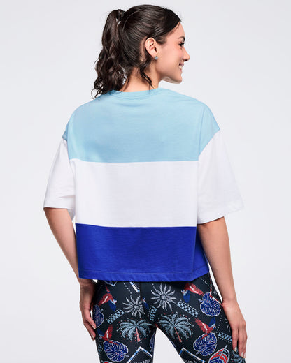 Back view of the light blue, white, and royal blue color-block oversized T-shirt on a female model, showing the wide sleeves and straight hem, paired with black tropical-print leggings.
