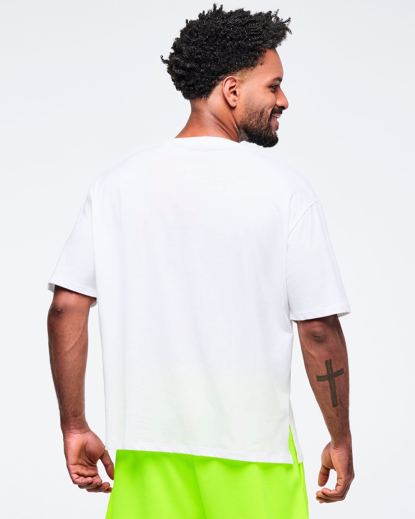 Back view of a white oversized Zumba T-shirt with a clean, solid back and relaxed fit. Male model pairs it with neon yellow shorts on a light gray studio background.