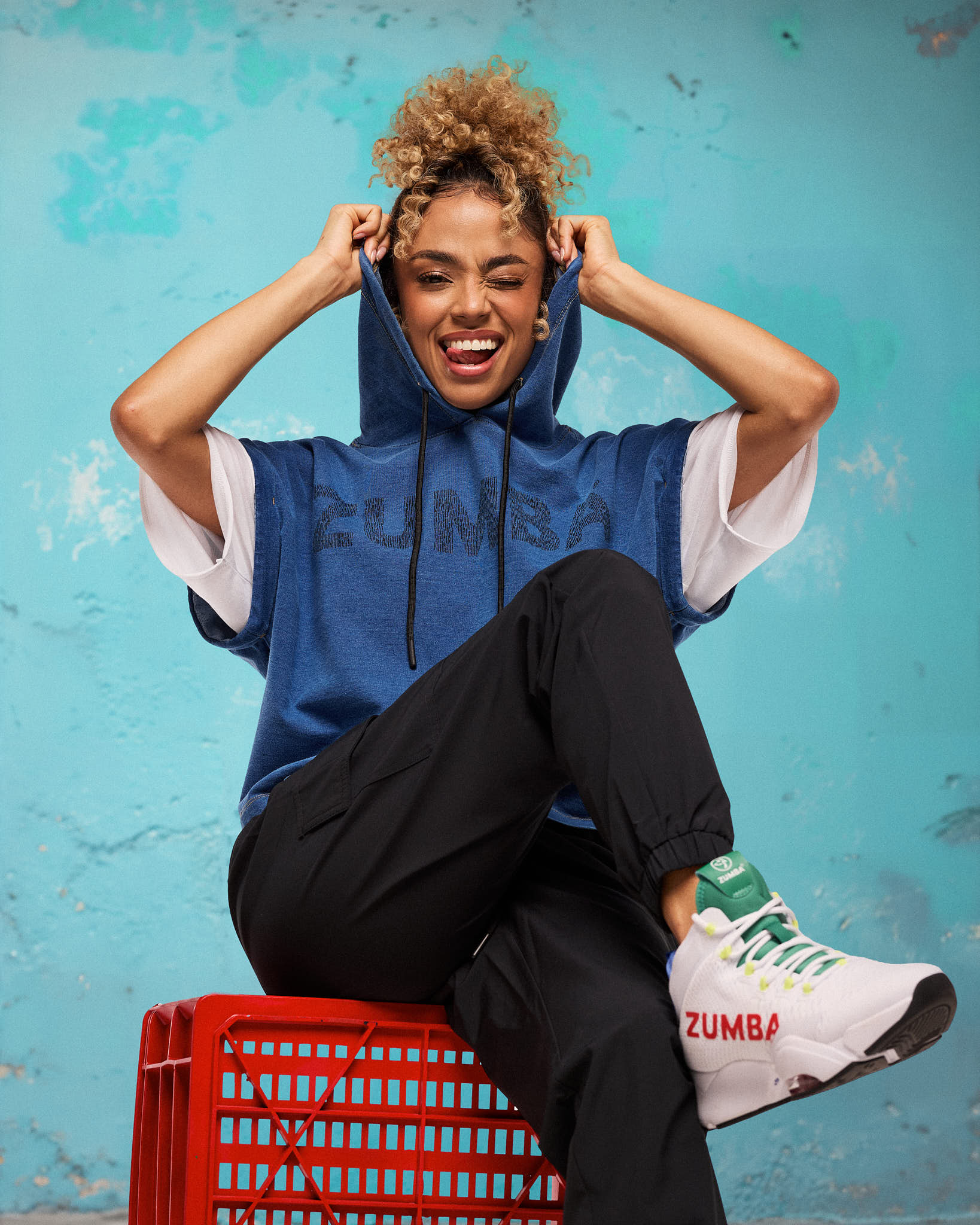 Navy, denim short-sleeve hooded sweatshirt with black drawstrings and tonal “ZUMBA” chest graphic; female model wears black jogger pants and white high-top sneakers with green accents, seated on a red crate against a turquoise wall.