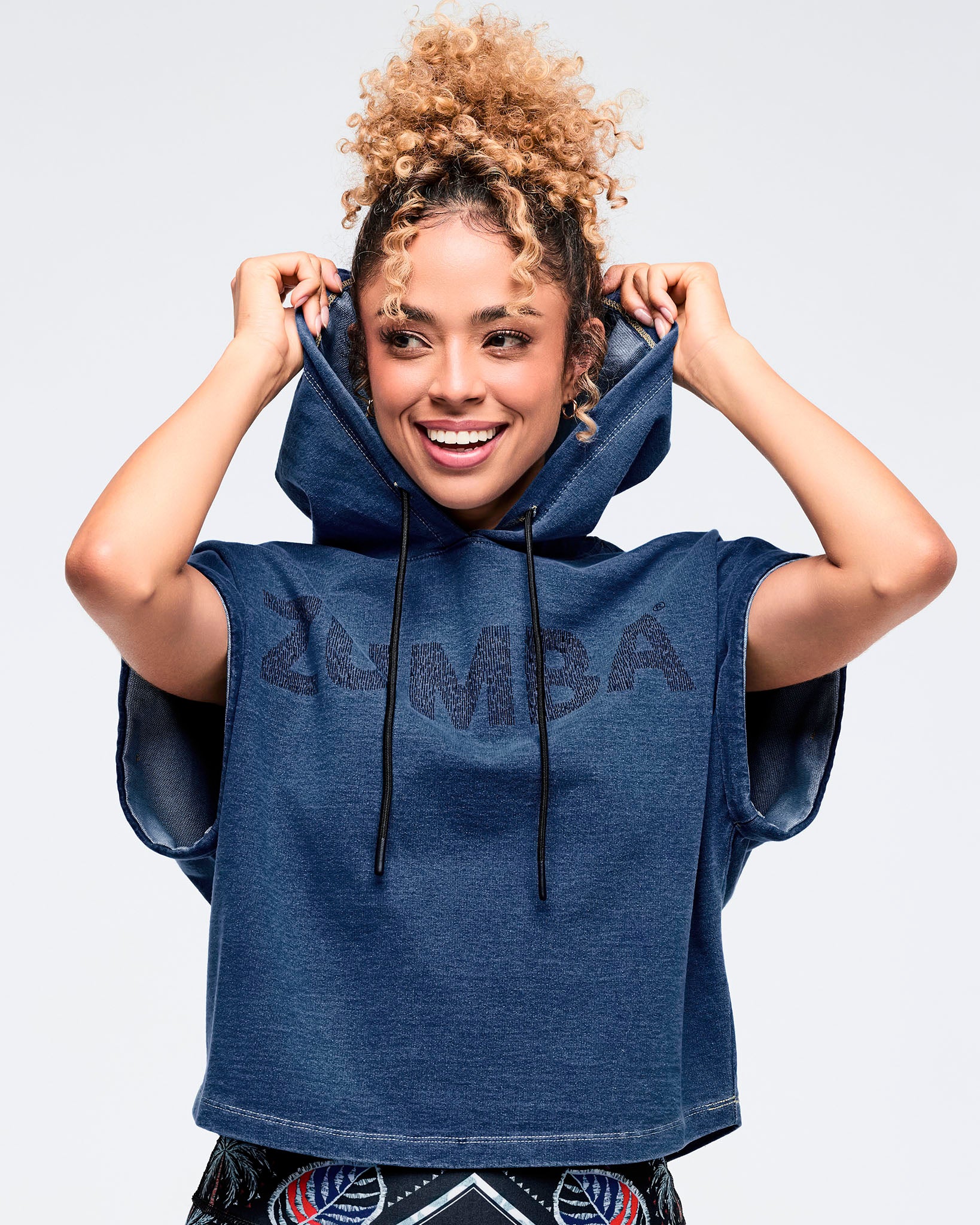 Close-up of a navy denim short-sleeve hoodie with black drawstrings and tonal “ZUMBA” logo across the chest on a female model against a white background.