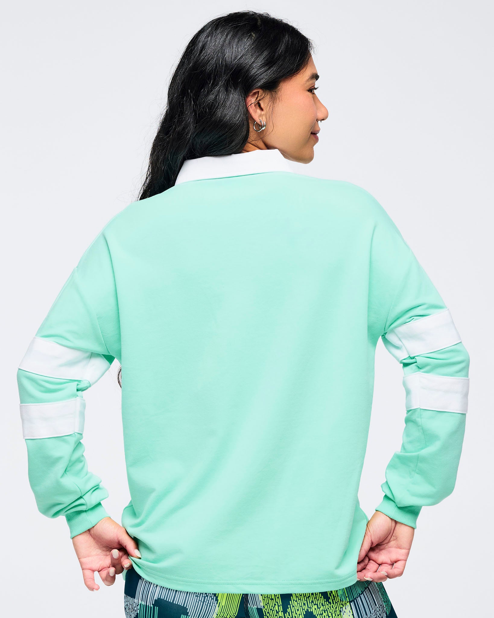Back view of the mint green rugby sweatshirt with white collar and double white sleeve stripes on a female model, paired with green abstract print bottoms, on a white background.