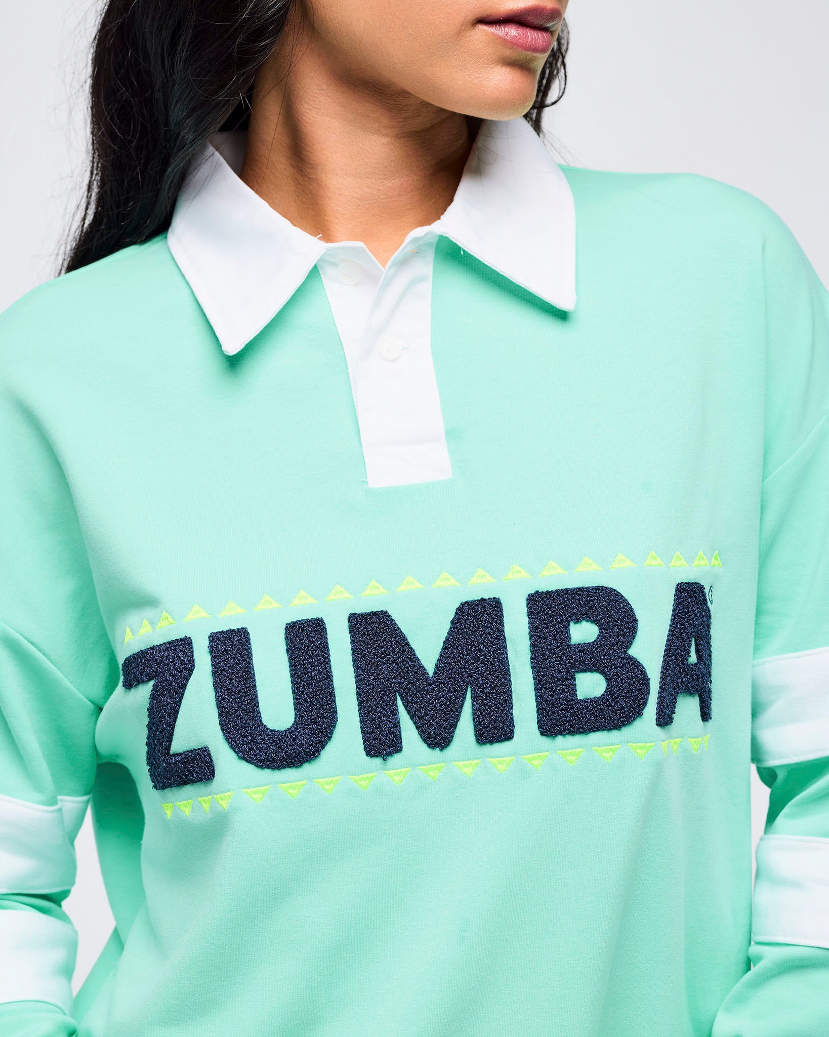 Close-up of the mint green rugby sweatshirt showing the white collar, button placket, and textured navy “ZUMBA” lettering on a female model.