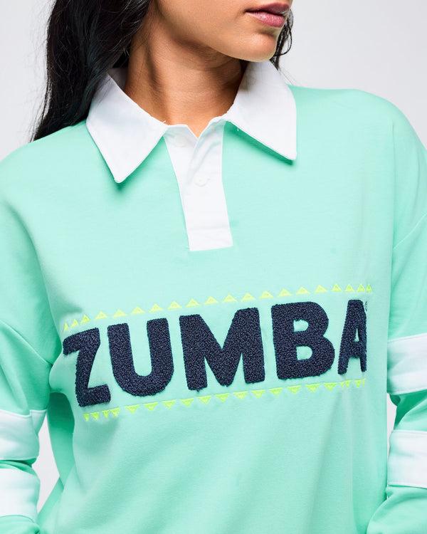 Close-up of the mint green rugby sweatshirt showing the white collar, button placket, and textured navy “ZUMBA” lettering on a female model.
