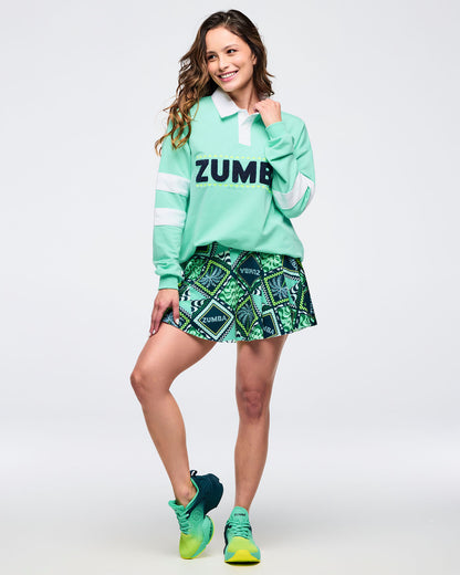 Mint green long-sleeve rugby sweatshirt with white collar and bold “ZUMBA” chest lettering; female model styles it with a green patterned flouncy skort and green sneakers on a white background.