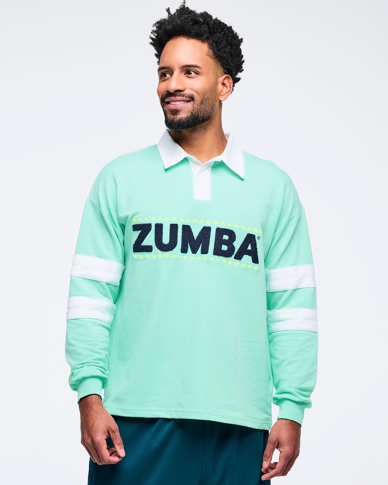 Front close-up of a mint green long-sleeve rugby sweatshirt featuring a white collar, white sleeve stripes, and large navy “ZUMBA” chest lettering on a male model against a white background.