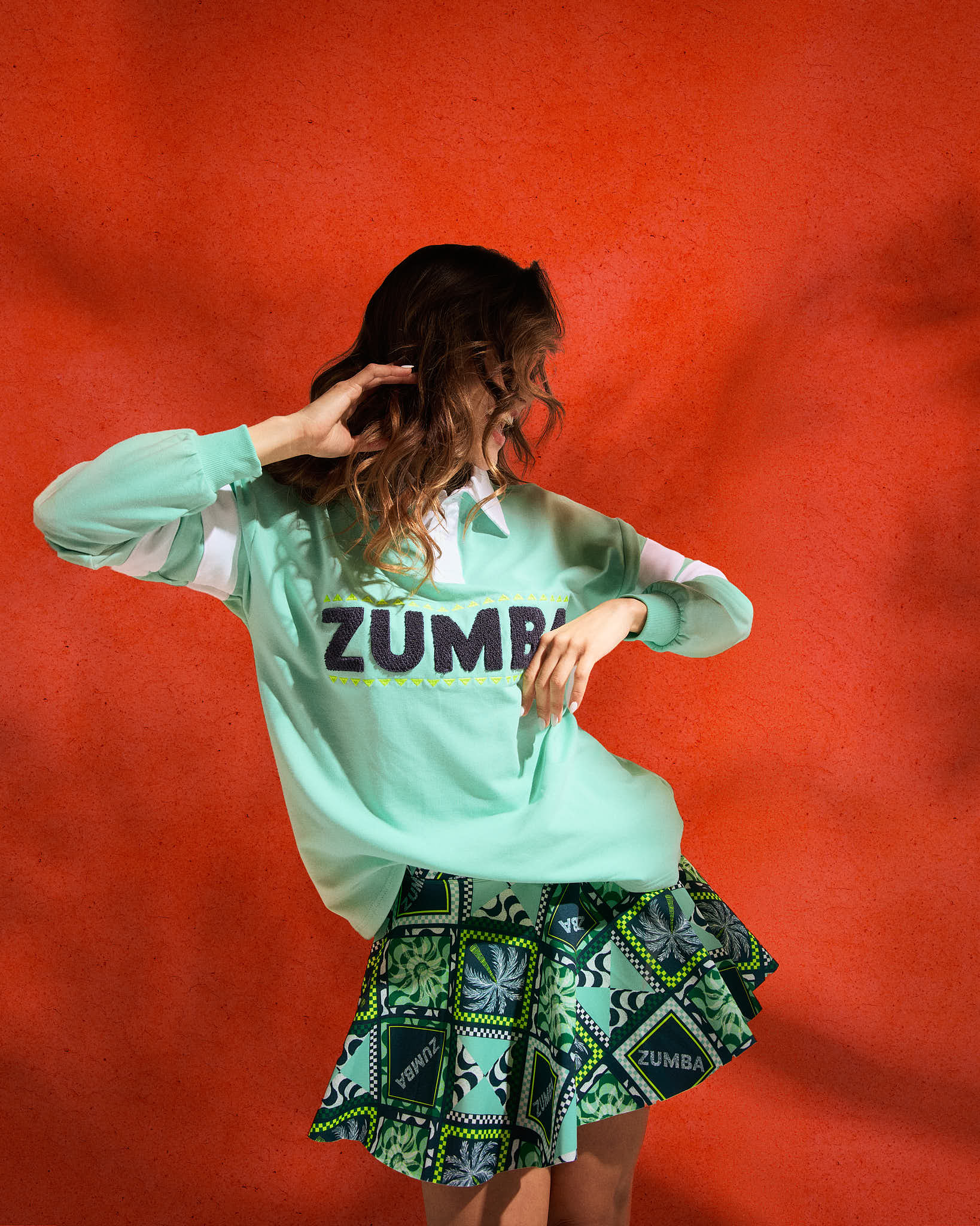 Green Zumba patchwork skort with flared hem and matching printed set peeking underneath. Female model wears a mint pullover sweatshirt with bold “ZUMBA” lettering, posing against a textured red wall.