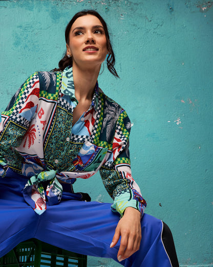 Multicolor patchwork Zumba button-up shirt with tropical prints and a front tie. Female model wears the long-sleeve collared shirt tied at the waist with royal blue track pants, posed against a teal textured wall.