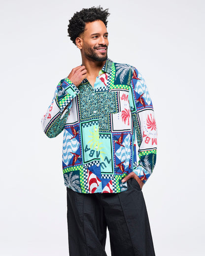Multicolor patchwork Zumba button-up shirt with tropical prints and checkerboard trim, styled open at the collar. Male model wears it with wide-leg black pants on a light gray studio background.