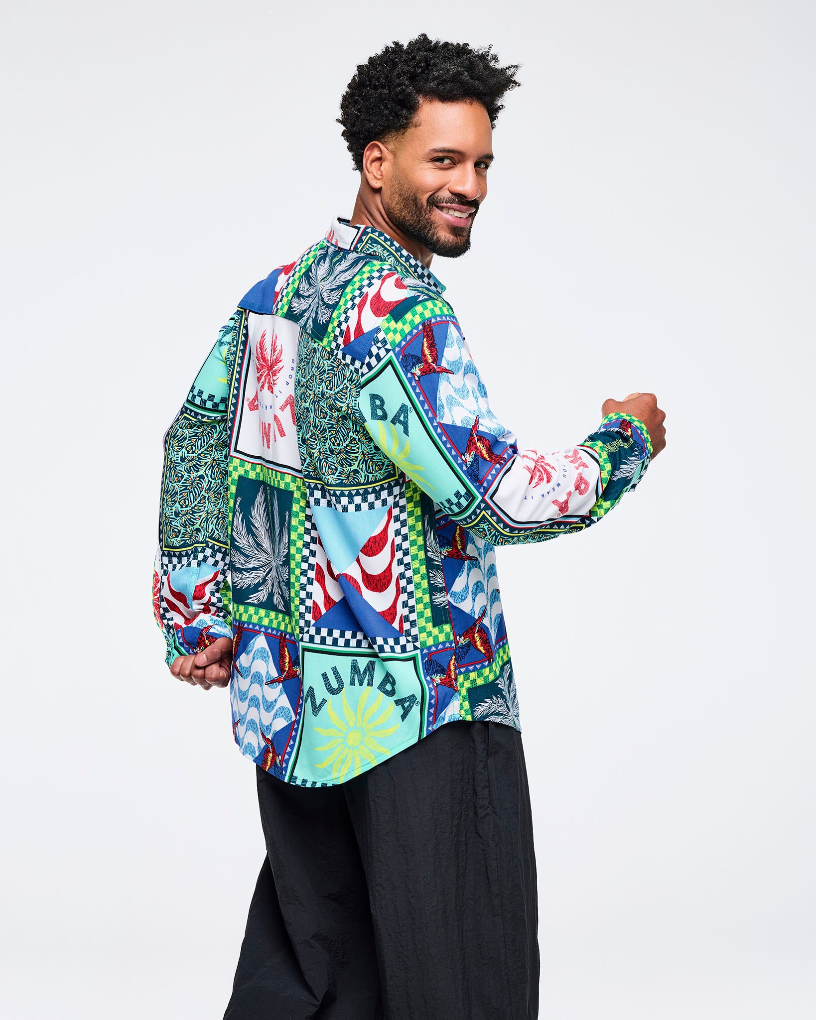 Multicolor patchwork Zumba button-up shirt featuring tropical and wave-print panels with checkerboard borders. Male model wears the long-sleeve collared shirt with wide-leg black pants on a light gray studio background.