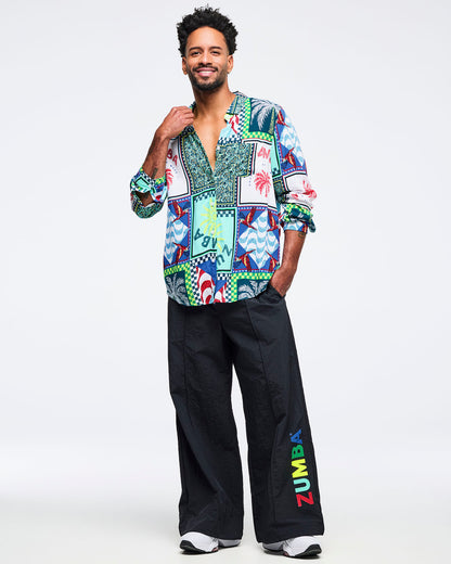 Full-body view of the multicolor patchwork Zumba button-up shirt worn with wide-leg black pants featuring vertical multicolor “ZUMBA” text on the leg and white sneakers. Male model poses on a light gray studio background.