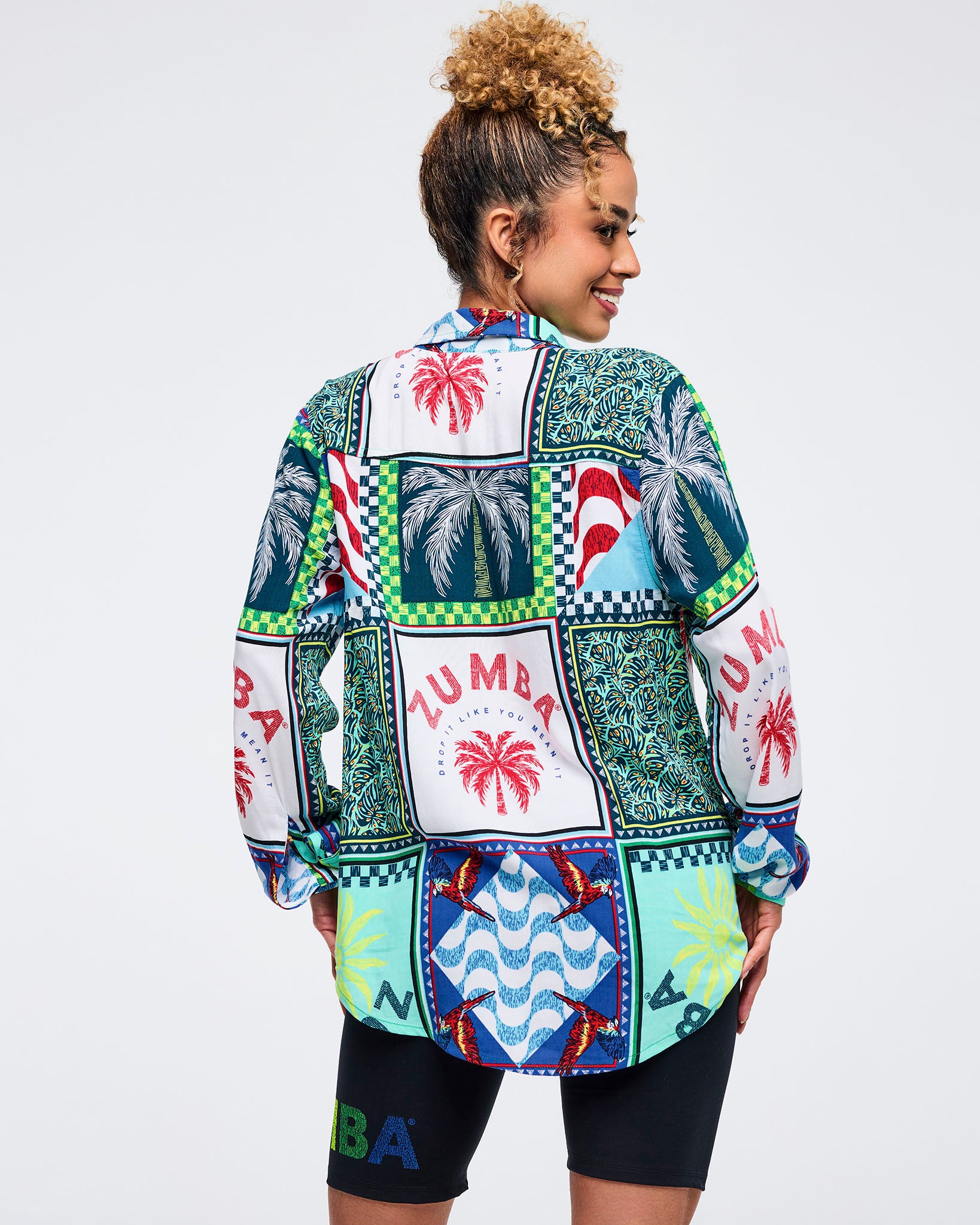 Back view of the multicolor patchwork Zumba button-up shirt showing large palm tree panels, parrot motifs, and “ZUMBA” graphics. Female model pairs it with black biker shorts on a light gray studio background.