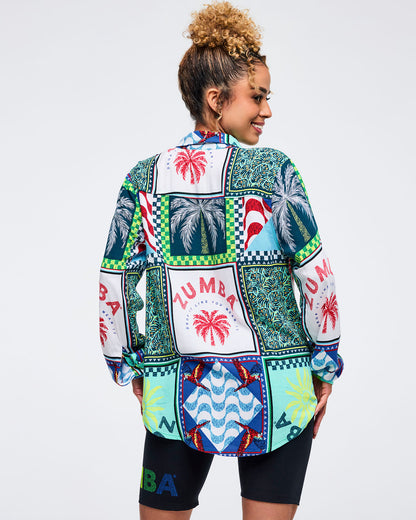 Back view of the multicolor patchwork Zumba button-up shirt showing large palm tree panels, parrot motifs, and “ZUMBA” graphics. Female model pairs it with black biker shorts on a light gray studio background.