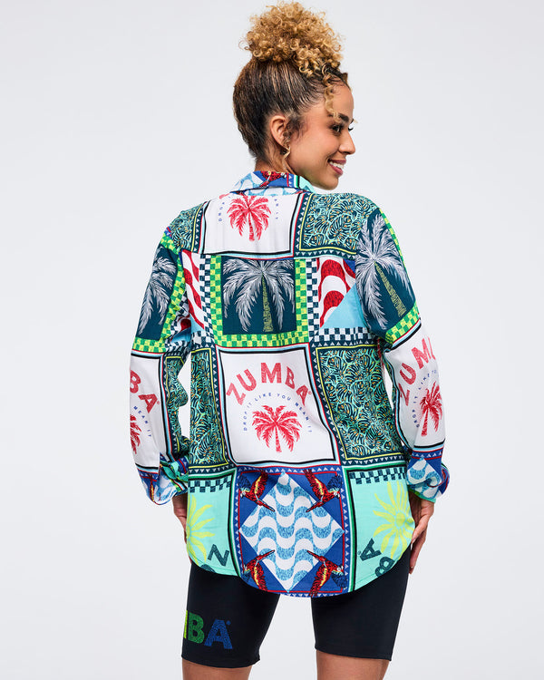 Back view of the multicolor patchwork Zumba button-up shirt showing large palm tree panels, parrot motifs, and “ZUMBA” graphics. Female model pairs it with black biker shorts on a light gray studio background.