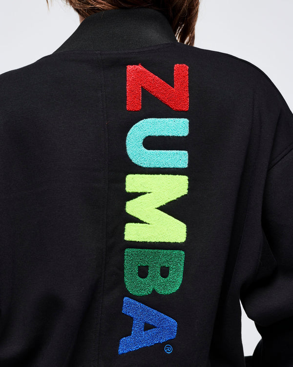 Close-up of the jacket back showing textured multicolor vertical “ZUMBA” lettering on black fabric with a ribbed collar detail.