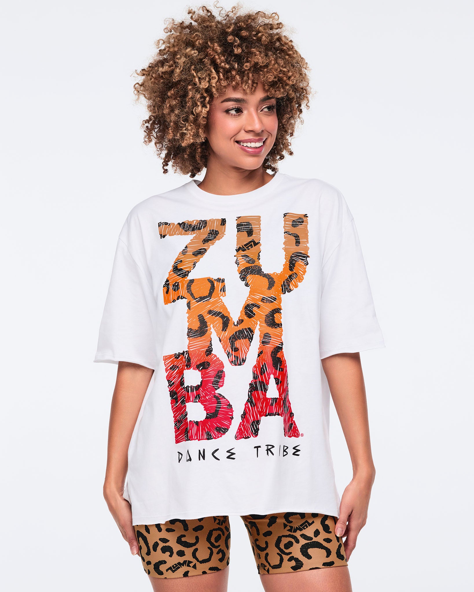 White oversized T-shirt with large orange and red leopard-print “Zumba Dance Tribe” graphic lettering, shown on a female model wearing matching leopard-print biker shorts.