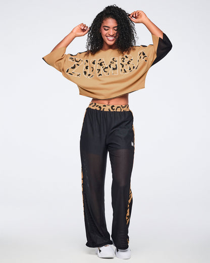 Tan top with oversized cheetah-print “Zumba” lettering, wide three-quarter sleeves, and black mesh sleeve panels, shown on a smiling female model wearing black mesh wide-leg pants with tan cheetah-print side stripes and white sneakers.