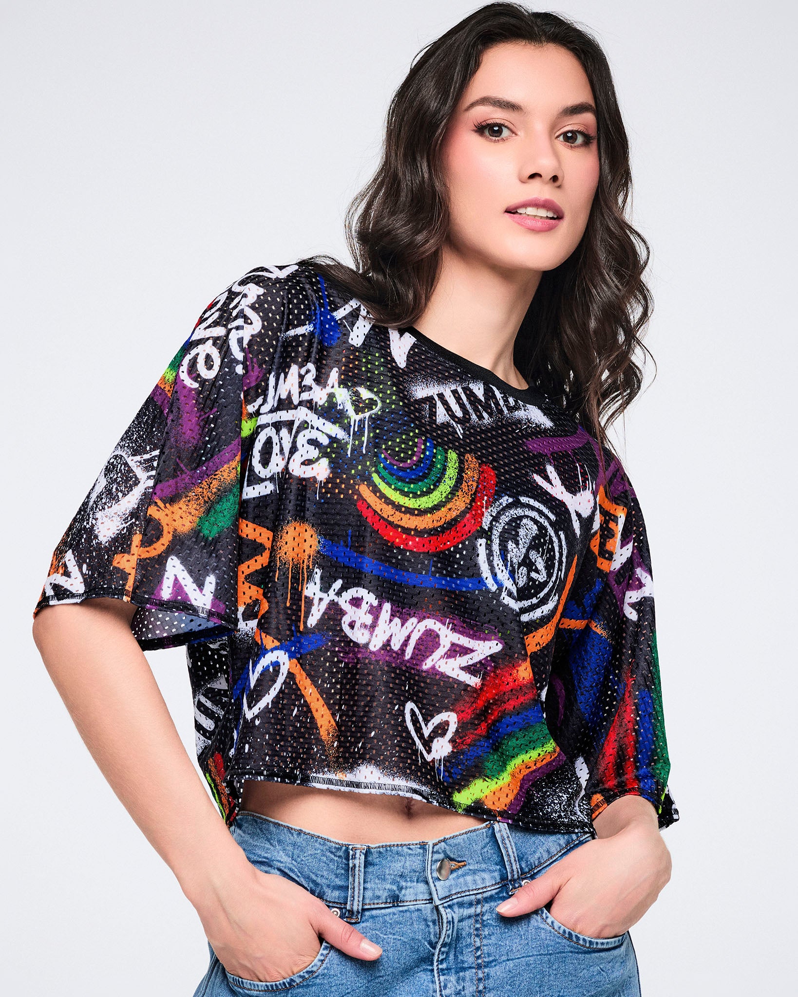 Female model wearing a black mesh crop top with colorful text and designs, paired with denim cargo pants on a white background.