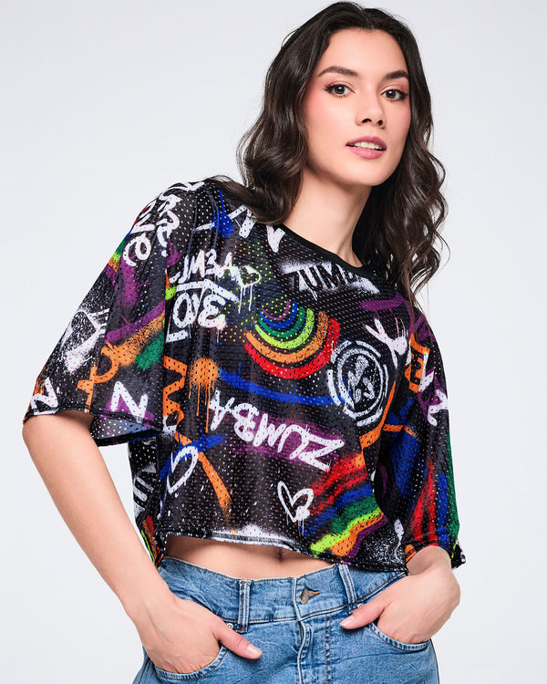Female model wearing a black mesh crop top with colorful text and designs, paired with denim cargo pants on a white background.