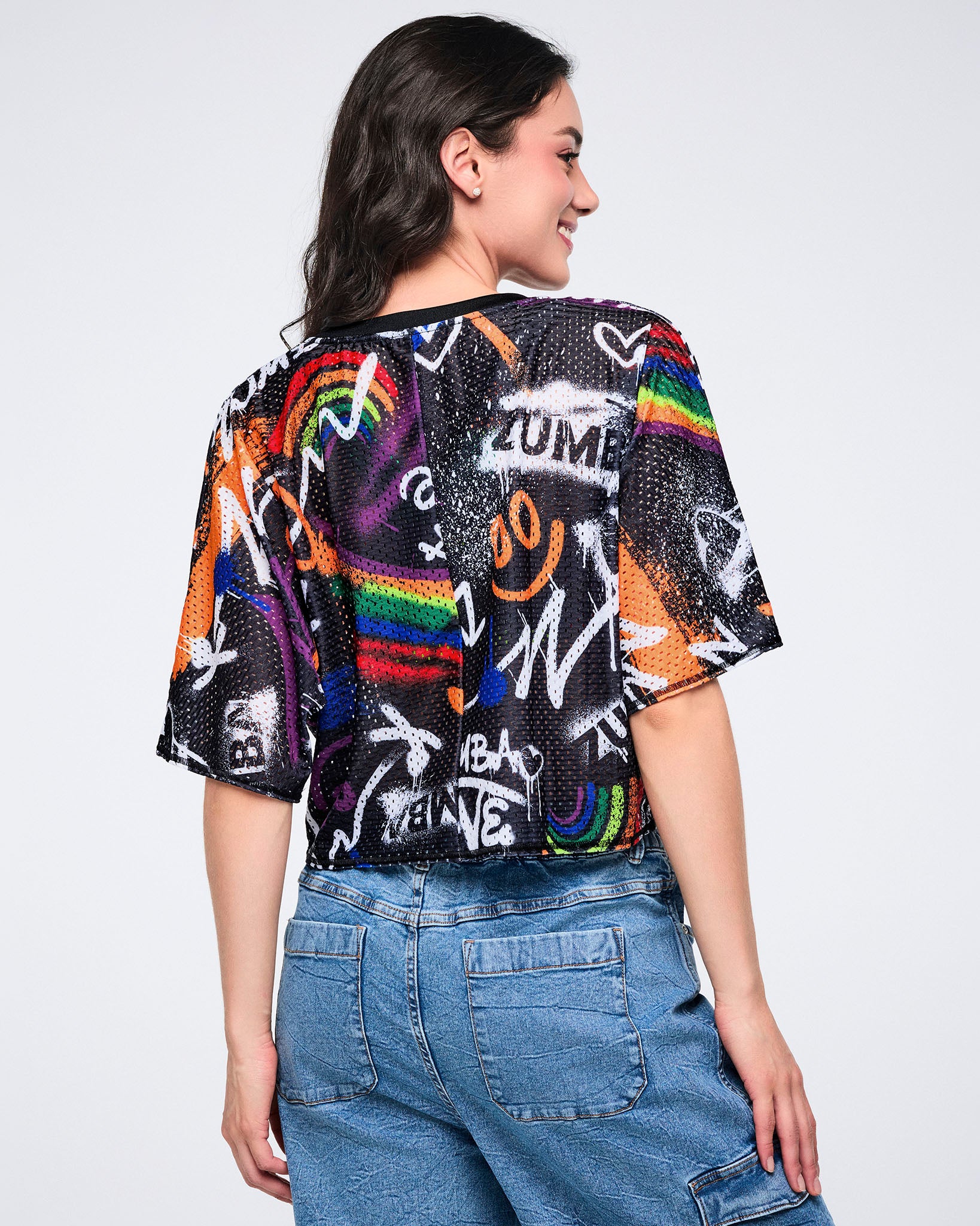 Female model, shown from behind, wearing a colorful graphic mesh crop top with text and rainbow designs paired with denim cargo pants on a white background.