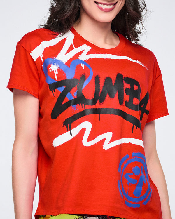 Female model wearing a red 'Zumba' shirt with colorful graphics on a white background.