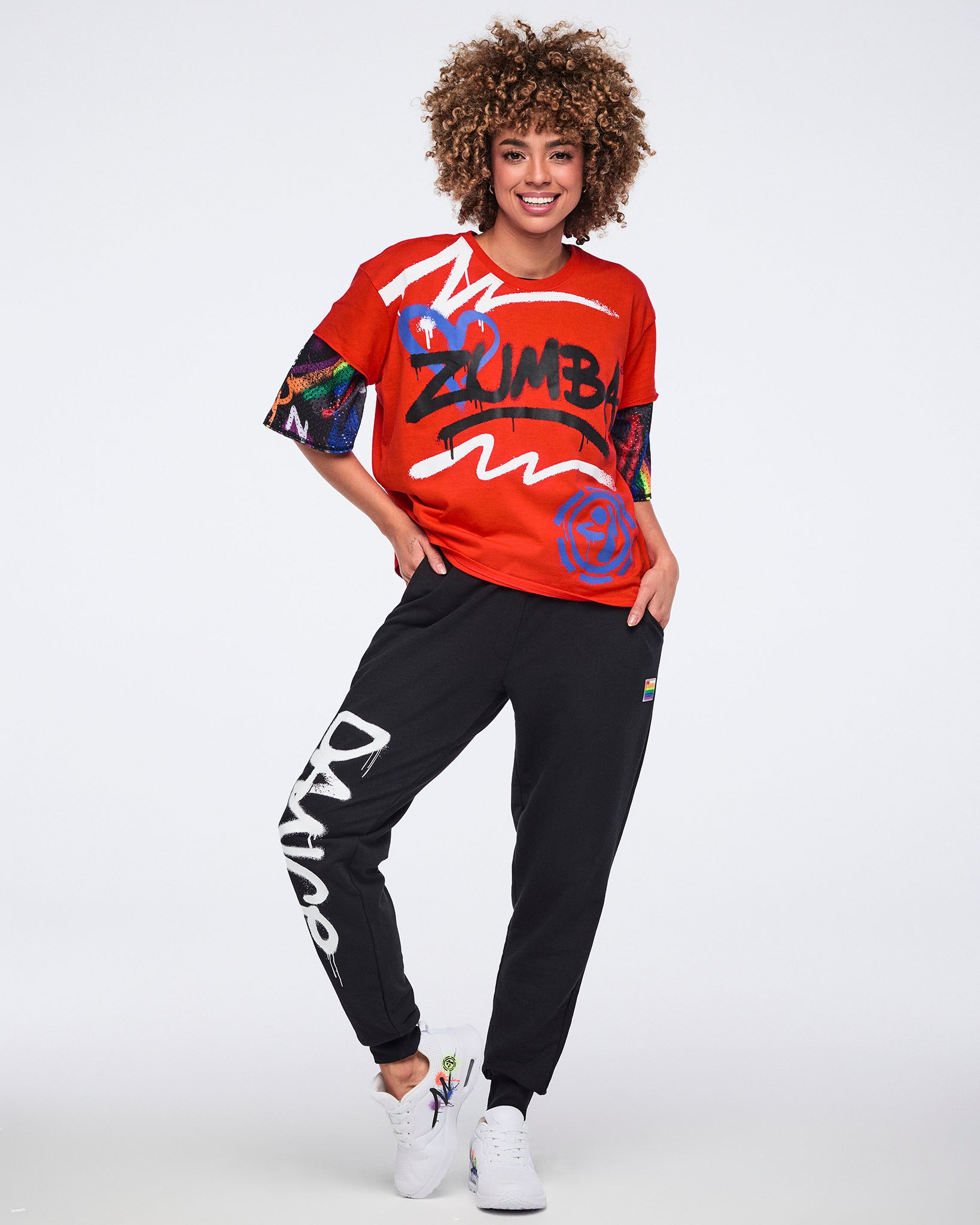 Female model wearing a red Zumba shirt and black pants with 'DANCE' down the right leg, standing against a white background.