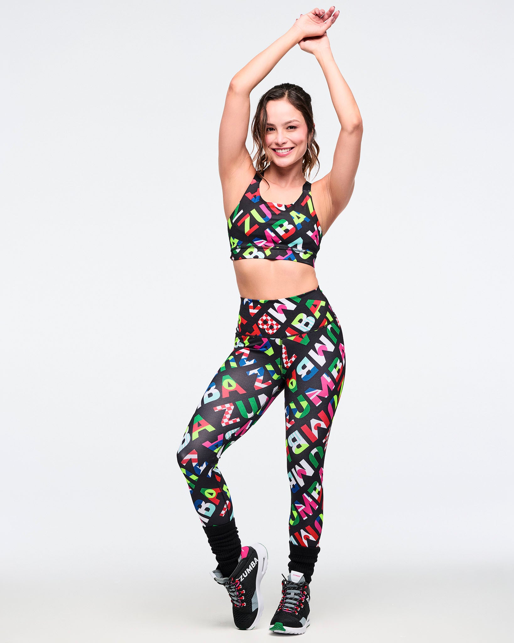 Full view of a female model wearing a colorful sports bra paired with matching leggings black and pink shoes. on a white background.