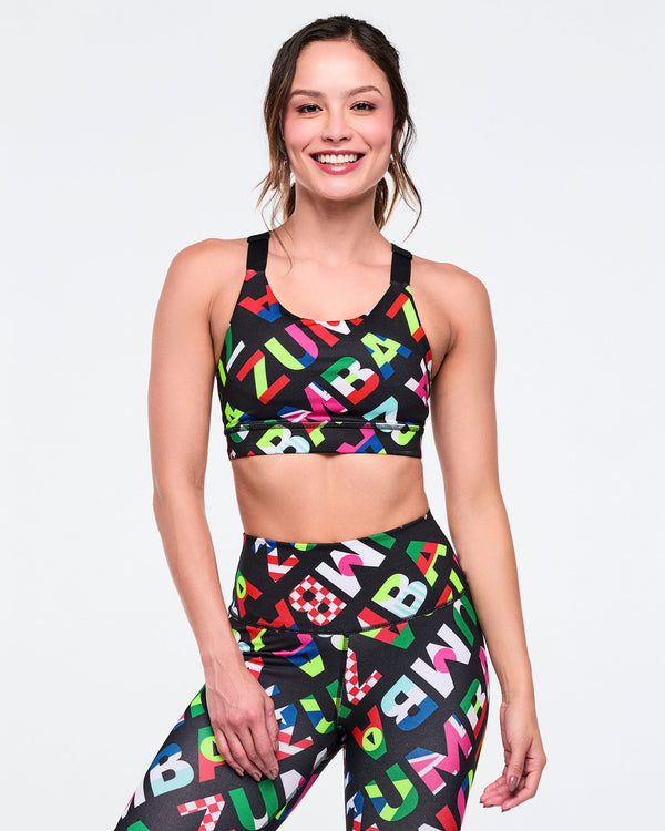 Female model wearing sports bra with colorful flag pattern on 'Zumba' letters paired with matching leggings on a white background.