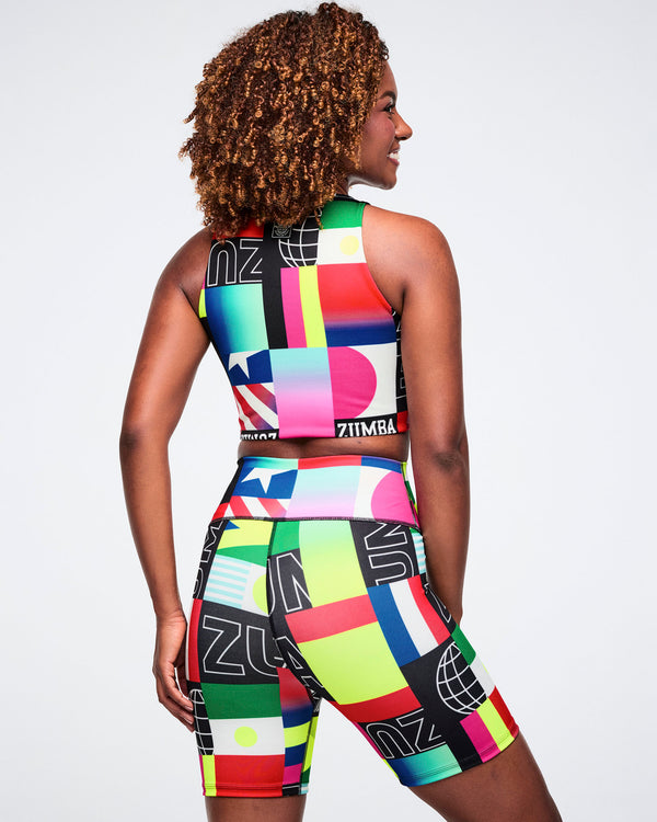 Back view of a female model wearing a colorful crop top and biker shorts with geometric patterns and brand logos on a plain background.