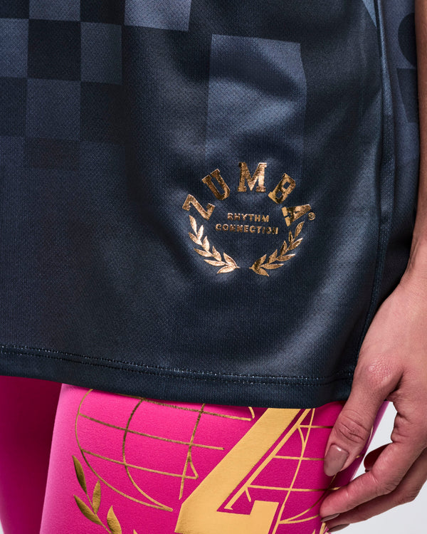 Close-up of a black top’s lower left side with a gold “Zumba Rhythm Connection” logo, paired with pink leggings with gold accents.