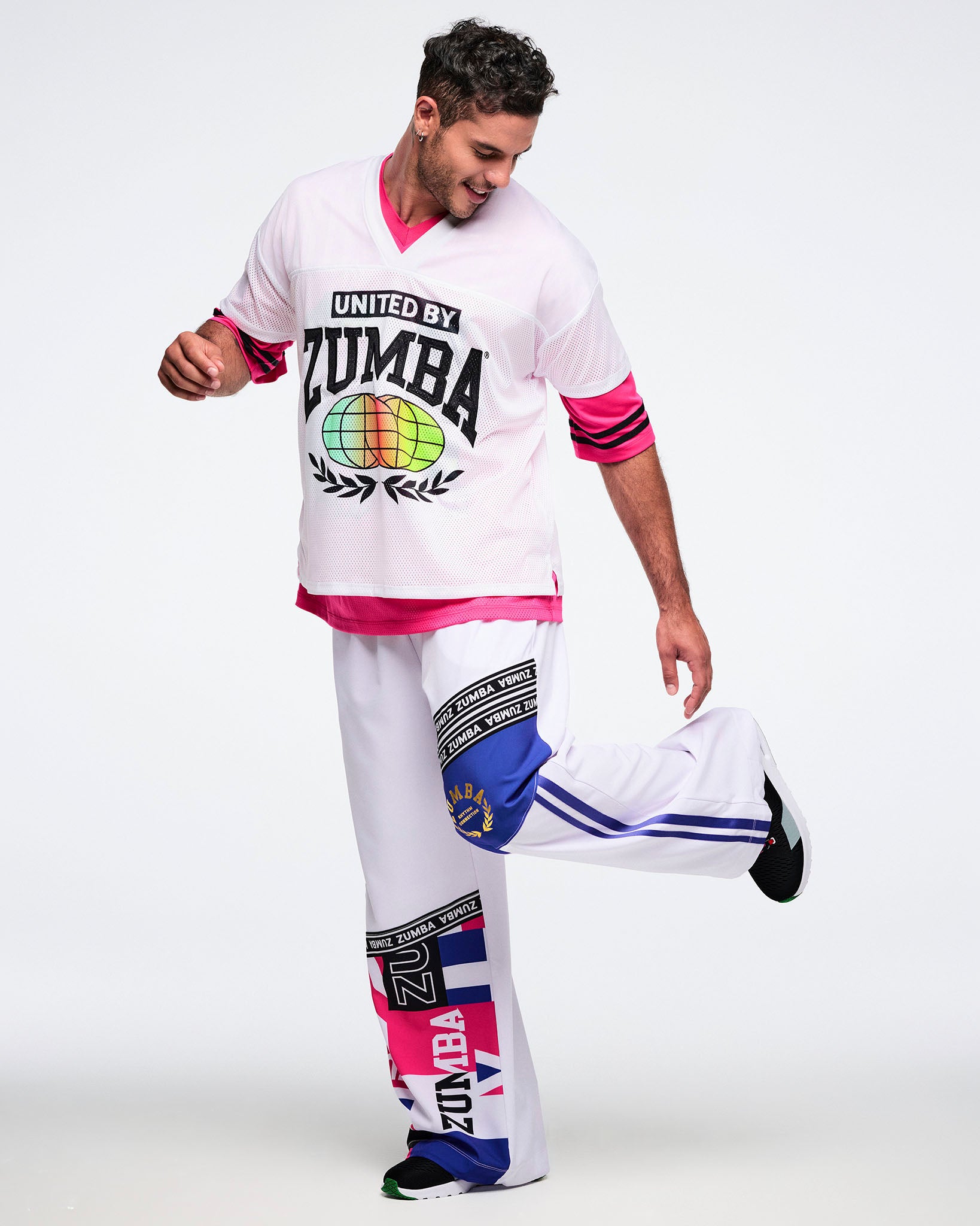 Male model wearing a white Zumba outfit - a white 'United by Zumba' top with a colorful globe design and matching white pants with blue, pink, black and white accents against a white background.