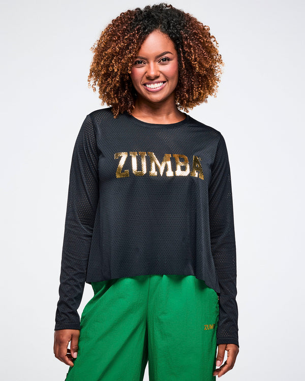 Female model wearing a black Zumba mesh shirt with gold 'ZUMBA' lettering in the center, paired with green pants on a white background.