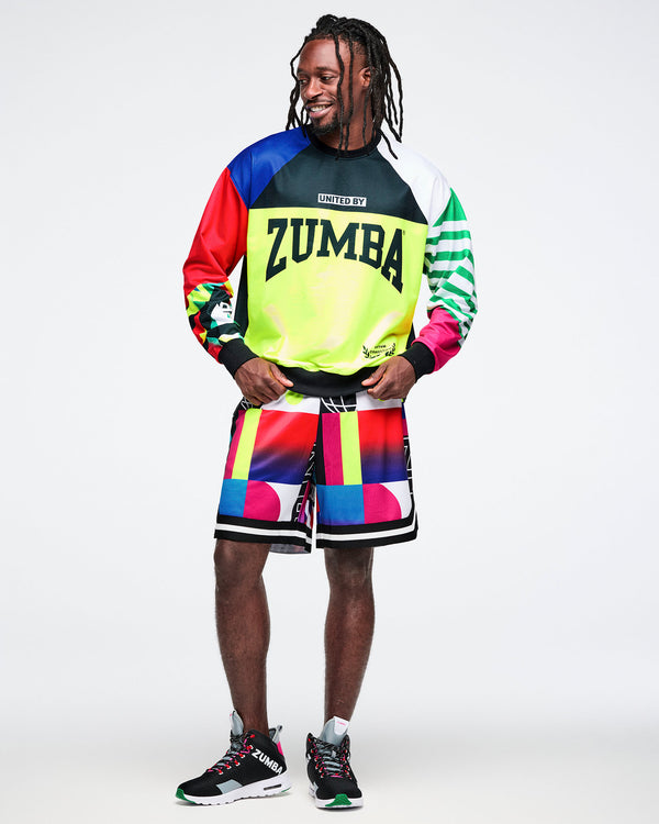 Full-body view of a male model in a colorful Zumba top, matching geometric-patterned shorts, and black-and-pink shoes, set against a white background.