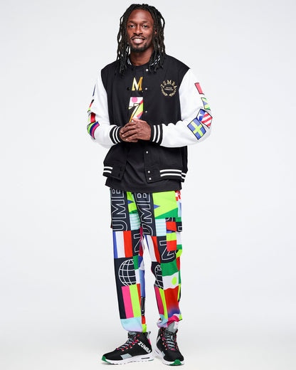 Male model in an unbuttoned colorful varsity jacket over a black Zumba tee, with matching multicolor flag pants on a white background.