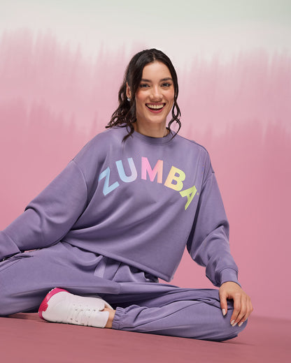 Female model wearing a purple crewneck sweatshirt with multicolor “ZUMBA” lettering, paired with matching purple jogger sweatpants and white sneakers with pink accents.