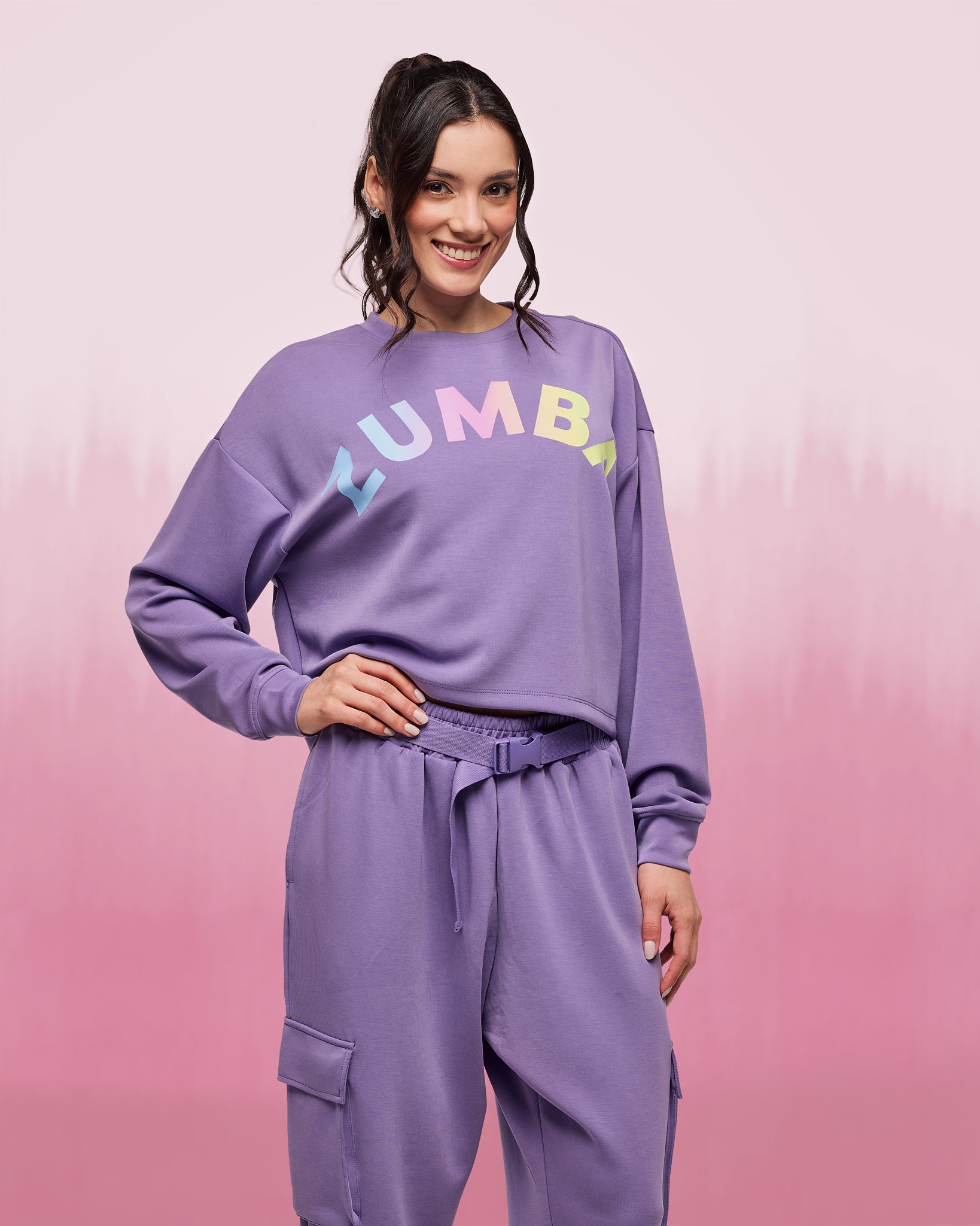 Female model wearing a purple cropped crewneck sweatshirt with pastel “ZUMBA” lettering across the chest, paired with matching purple high-waisted cargo jogger pants featuring flap pockets, a drawstring waist, and a tonal web belt with buckle.